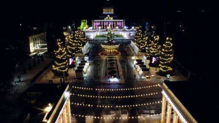Foto de Paisaje vista gran edificio, árboles de Navidad, niños girando carrusel y pista de patinaje sobre hielo al aire libre. Iluminación brillante guirnaldas decoraciones Año Nuevo Navidad - Imagen libre de derechos
