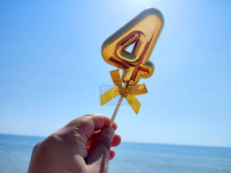 Person holding stick with a shiny golden number 4 four in his hand on backdrop of the waves of the blue sea and blue sky on a sunny summer day. Concept holiday birthday party celebrating