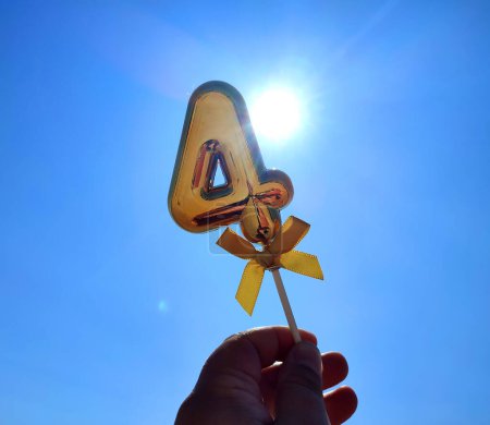 Person holding stick with a shiny golden number 4 four in his hand on backdrop of the waves of blue sky and shining sun on a sunny summer day. Concept holiday birthday party celebrating