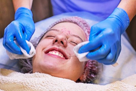 Photo for Happy girl teenager on the procedure at the beautician, cleaning her face. beautician wipes face with special wipes after mask - Royalty Free Image