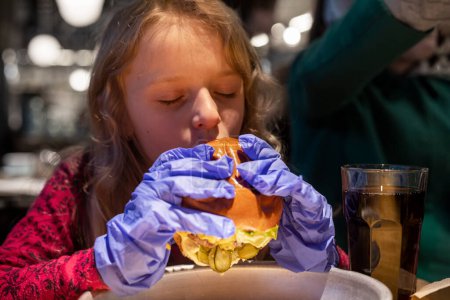 Photo for Happy little girl with closed eyes bites off a hamburger and holds it with both hands in disposable gloves - Royalty Free Image