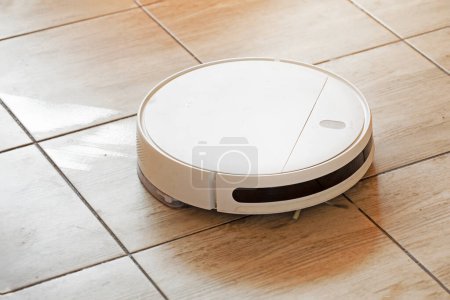 Foto de Aspiradora robot limpia y lava los pisos en el suelo de baldosas en la sala de estar iluminada por el sol - Imagen libre de derechos
