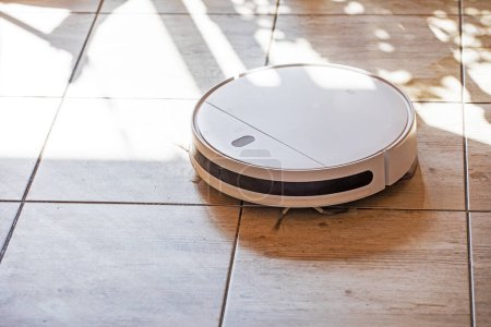 Foto de Aspiradora robot limpia el suelo de baldosas en la sala de estar iluminada por el sol, horizontal - Imagen libre de derechos