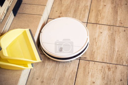 aspiradora robot limpia en un suelo de baldosas en una sala de estar iluminada por el sol junto a las ollas amarillas para la limpieza, horizontal