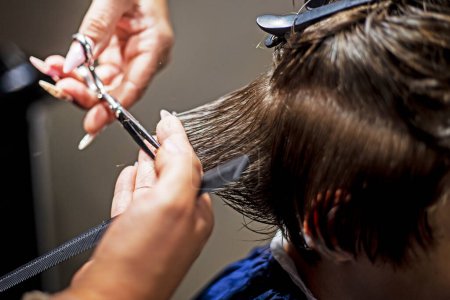 Téléchargez les photos : Coiffeur crée une coupe de cheveux à la mode pour un garçon ténager aux cheveux blonds foncés - en image libre de droit