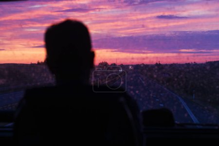 Téléchargez les photos : Silhouette sombre floue regarde à travers le verre sale du bus au lever du soleil avec des nuages sur la route. Voyage - en image libre de droit