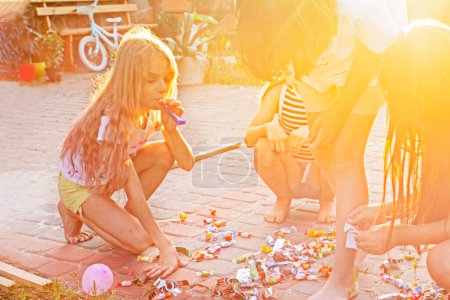 Téléchargez les photos : Fille heureuse avec les cheveux longs soufflant une paille à une fête d'enfants éclairée par le soleil - en image libre de droit