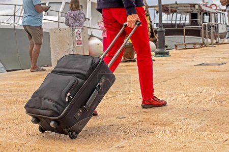 Téléchargez les photos : Fille en pantalon rouge porte une valise noire sur roues sur la jetée lors de l'embarquement sur un navire. Vacances et voyages - en image libre de droit