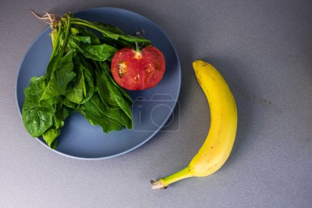 Téléchargez les photos : Banane se trouve sur le comptoir à côté d'une assiette d'épinards et une demi-tomate rouge - en image libre de droit