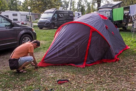 Photo for Man squatting puts up a tent at a campsite in the evening. travel and vacation by car - Royalty Free Image