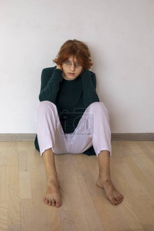 Foto de Feliz chica adolescente con estilo con gafas y pelo de anime rojo se sienta en el suelo de parquet. Psicología de la edad - Imagen libre de derechos