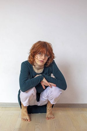 Téléchargez les photos : Adolescente avec des cheveux roux et des lunettes squats près du mur. Psychologie et complexes d'adolescents - en image libre de droit