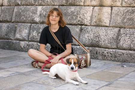 happy girl in glasses sits with a Jack Russell Terrier dog on a sunny day on the street