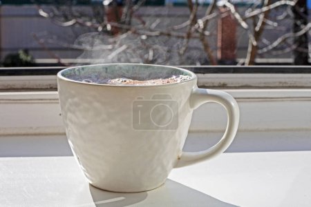 white cup of hot drink with tea or coffee stands on the sill near the window. Day off
