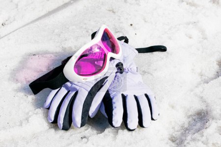 Téléchargez les photos : Masque de ski rose pour enfants avec gants de ski se trouvent sur une pente enneigée humide par une journée ensoleillée. - en image libre de droit