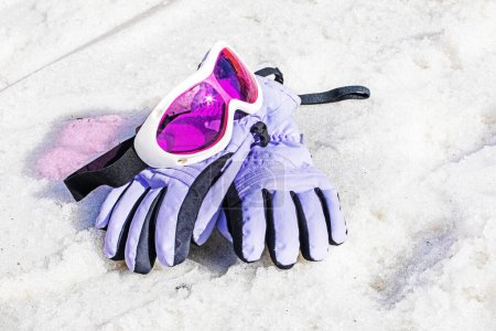 pink children's snow mask with snow gloves lie on a wet snowy slope on a sunny day. Winter snow season. Active holiday with family