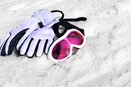 pink children's ski mask with ski gloves lie on a wet snowy slope on a sunny day.