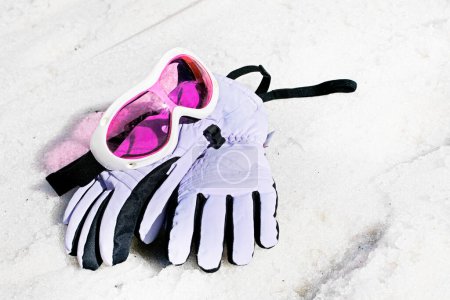 Pinkfarbene Kinderskimaske mit Skihandschuhen liegt an einem sonnigen Tag auf einem nassen, schneebedeckten Hang.
