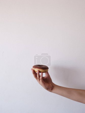 Süßes in sanfter Hand. Schokoladenglasierter Donut in der Hand vor einem schlichten, aufgeräumten, hellen Hintergrund
