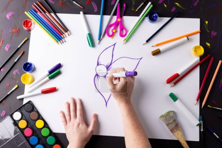 Téléchargez les photos : Couvre-chef des mains dessinant des fleurs violettes sur papier, avec des matériaux d'art sur le dessus de table. Art, créativité, mois artisanal et concept de passe-temps. - en image libre de droit