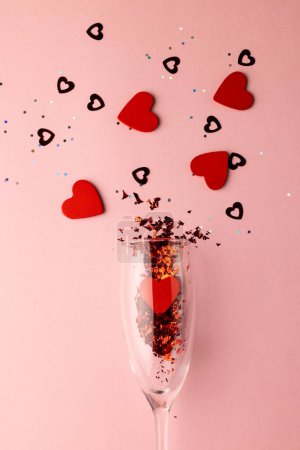 Photo for Vertical of champagne glass spilling red heart shapes on pale pink background with copy space. Valentine's day, love, romance and celebration concept. - Royalty Free Image