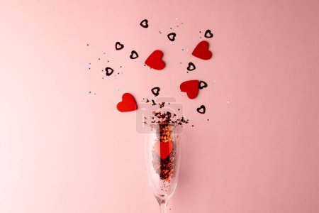 Téléchargez les photos : Verre à champagne renversant des formes de coeur rouge et des confettis sur fond rose pâle avec espace de copie. Saint Valentin, amour, romance et concept de célébration. - en image libre de droit