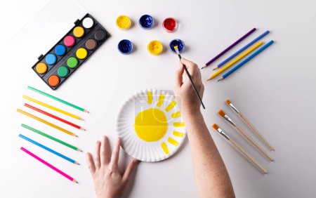 Photo for Overhead of hands painting with yellow paint on paper plate, with art materials on table top. Art, creativity, craft month and hobbies concept. - Royalty Free Image