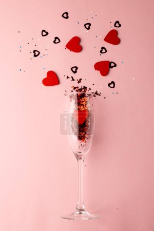 Foto de Vertical de copa de champán derramando formas de corazón rojo sobre fondo rosa pálido con espacio de copia. Día de San Valentín, amor, romance y concepto de celebración. - Imagen libre de derechos