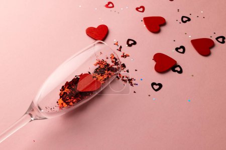 Téléchargez les photos : Verre à champagne renversant des formes de coeur rouge et des confettis sur fond rose pâle avec espace de copie. Saint Valentin, amour, romance et concept de célébration. - en image libre de droit