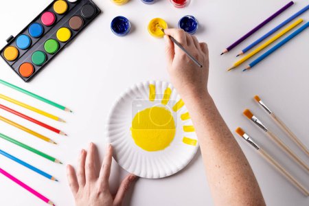 Photo for Overhead of hands painting with yellow paint on paper plate, with art materials on table top. Art, creativity, craft month and hobbies concept. - Royalty Free Image