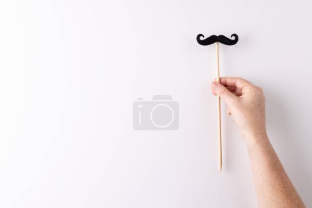 Photo for Hand holding black false moustache on stick, on white background with copy space. Father's day, moustache day, dressing up, party props, fun and celebration concept. - Royalty Free Image