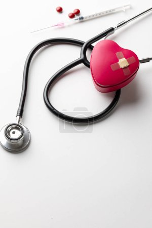 Photo for Vertical of stethoscope, heart with sticking plasters, syringe and pills, on white with copy space. Medical services, healthcare and health awareness concept. - Royalty Free Image