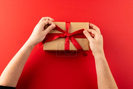Foto de Sobrecarga de manos atando regalo en papel marrón con cinta roja, sobre fondo rojo con espacio para copiar. Día de San Valentín, amor, romance y concepto de celebración. - Imagen libre de derechos