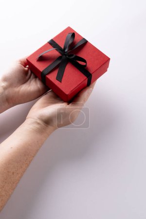 Téléchargez les photos : Vertical des mains tenant boîte cadeau rouge attaché avec ruban noir, sur fond blanc avec espace de copie. Saint Valentin, anniversaire, amour, romance et concept de célébration. - en image libre de droit