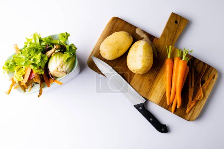 Photo for Overhead of knife and vegetables on chopping board with vegetable waste in kitchen composting bin. On white background with copy space. Ecology, recycling, care and nature concept. - Royalty Free Image