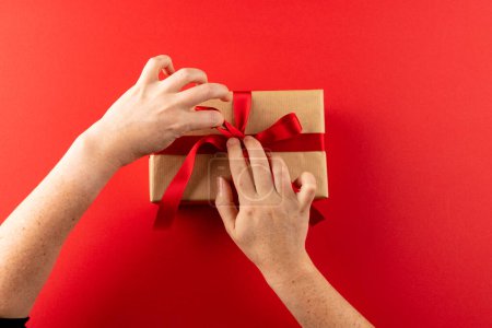 Téléchargez les photos : Couvre-chef des mains attachant cadeau en papier brun avec ruban rouge, sur fond rouge avec espace de copie. Saint Valentin, amour, romance et concept de célébration. - en image libre de droit