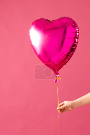Photo for Vertical of hand holding shiny pink heart shaped balloon on pink background with copy space. Valentine's day, love, romance and celebration concept. - Royalty Free Image