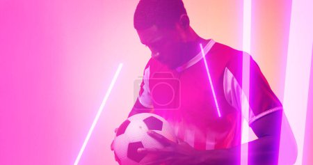Téléchargez les photos : Des lignes éclairées sur un joueur afro-américain sérieux tenant un ballon de football sur fond néon. Copier l'espace, composite, sport, compétition, jeu, match, néon et concept abstrait. - en image libre de droit