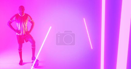 Photo for African american male player with leg on soccer ball and hands on hip standing by illuminated lines. Copy space, composite, sport, competition, shape, playing, match, neon and abstract concept. - Royalty Free Image