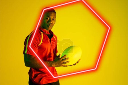 Photo for Hexagon neon over confident african american young rugby player holding ball on yellow background. Digital composite, sport, rugby, athlete, competitive sport, skill, copy space, portrait. - Royalty Free Image
