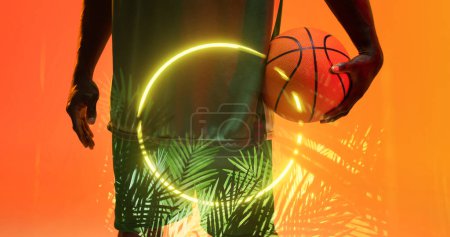 Téléchargez les photos : Milieu du joueur afro-américain de basket-ball tenant le ballon par cercle éclairé et plantes. Espace de copie, composite, main, sport, compétition, illustration, illuminé, nature, forme et abstrait. - en image libre de droit