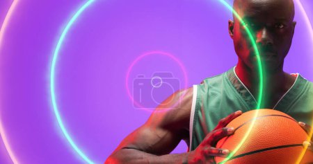 Téléchargez les photos : Portrait d'un joueur de basket américain chauve et sérieux tenant le ballon sur des cercles multicolores. Violet, espace de copie, composite, illuminé, sport, compétition, illustration, forme et abstrait. - en image libre de droit