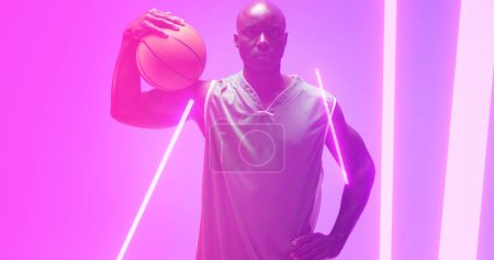 Téléchargez les photos : Composite d'afro-américain chauve joueur masculin tenant le basket-ball devrait par des lumières incandescentes. Rose, espace de copie, sérieux, illuminé, sport, compétition, illustration, forme et concept abstrait. - en image libre de droit