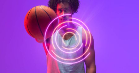 Téléchargez les photos : Composite de motif circulaire éclairé sur le portrait d'un joueur de basket biracial tenant un ballon. Espace de copie, violet, sport, compétition, illustration, éclat, forme et concept abstrait. - en image libre de droit