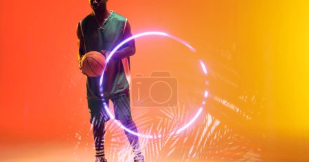 Téléchargez les photos : Milieu du joueur afro-américain de basket-ball tenant le ballon par cercle lumineux et des plantes. Espace de copie, composite, sport, compétition, illustration, illuminé, nature, forme et concept abstrait. - en image libre de droit