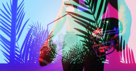 Foto de Midsection de jugador de fútbol americano con pelota y casco de pie por las plantas de color rosa y azul. Compuesto, mano, deporte, competición, ilustración, brillo, naturaleza y concepto abstracto. - Imagen libre de derechos