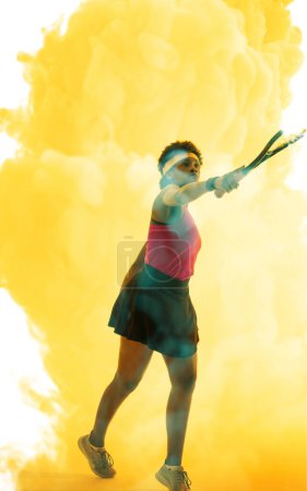 Photo for Full length of african american female player hitting with racket over yellow smoky background. Copy space, tennis, composite, sport, competition, playing, match and abstract concept. - Royalty Free Image