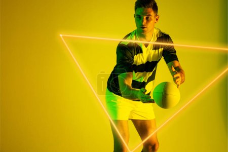 Foto de Jugador de rugby masculino caucásico atrapando pelota delante del triángulo iluminado sobre fondo amarillo. Copiar espacio, compuesto, deporte, forma, competencia, juego, partido y concepto abstracto. - Imagen libre de derechos