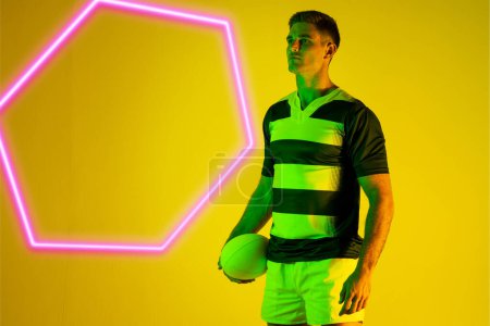 Photo for Caucasian male rugby player with ball standing by illuminated hexagon over yellow background. Copy space, composite, sport, shape, competition, playing, match and abstract concept. - Royalty Free Image