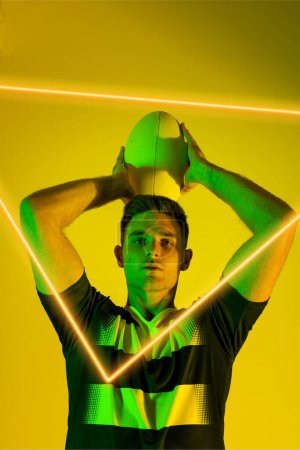 Foto de Retrato de jugador de rugby masculino caucásico lanzando pelota por triángulo iluminado sobre fondo amarillo. Copiar espacio, compuesto, deporte, forma, competencia, juego, partido y concepto abstracto. - Imagen libre de derechos